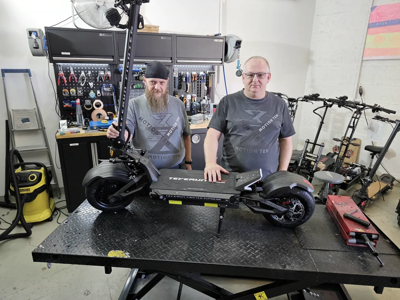Robert Hölzl und Oliver Köhler hinter einem Scooter auf der Hebebühne, dahinter die Werkstatt