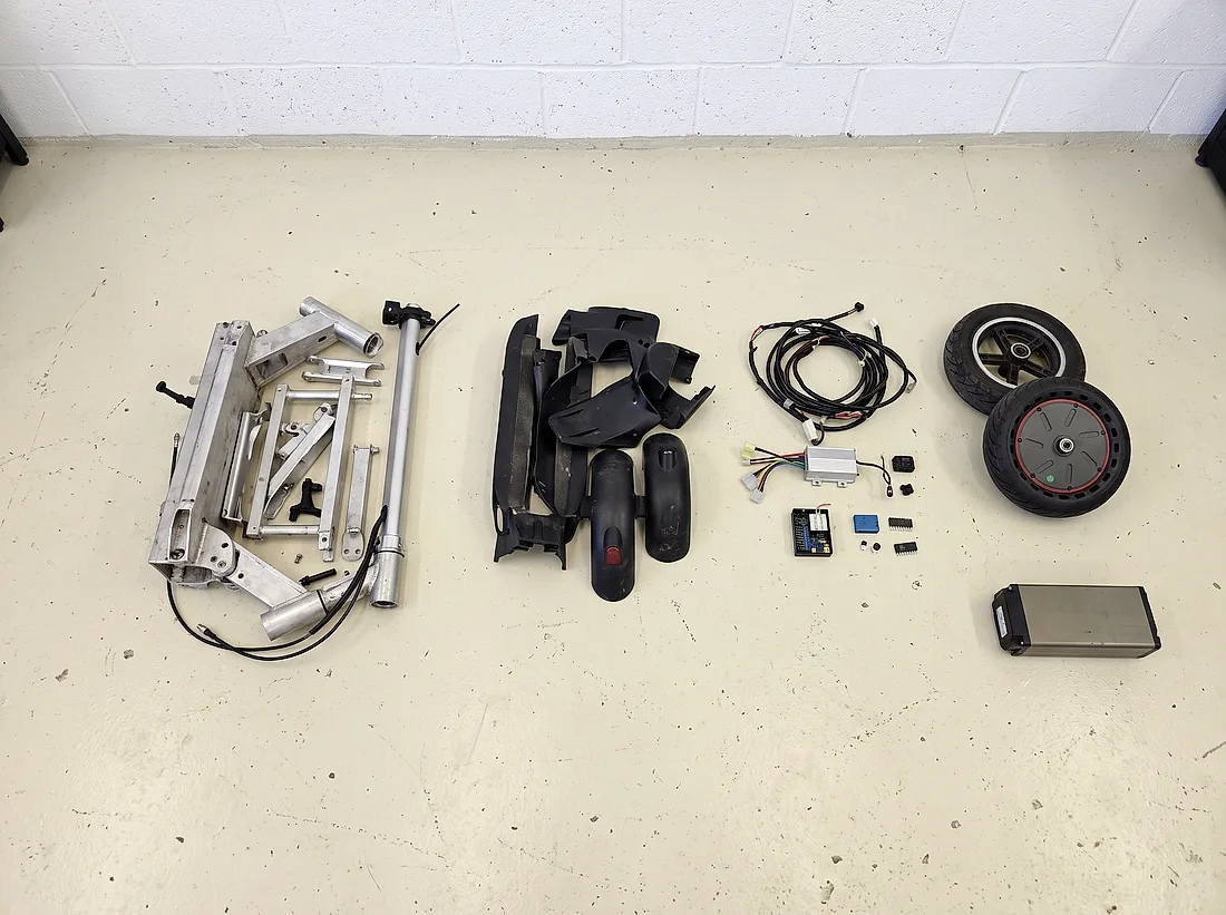 Zerlegter Scooter am Werkstattboden, die Materialien nach Gruppen getrennt ausgelegt: Aluminiumrahmen, Kunststoffverkleidung, Kabelbaum mit Elektronik, Räder, und abgesetzt davon der Akku