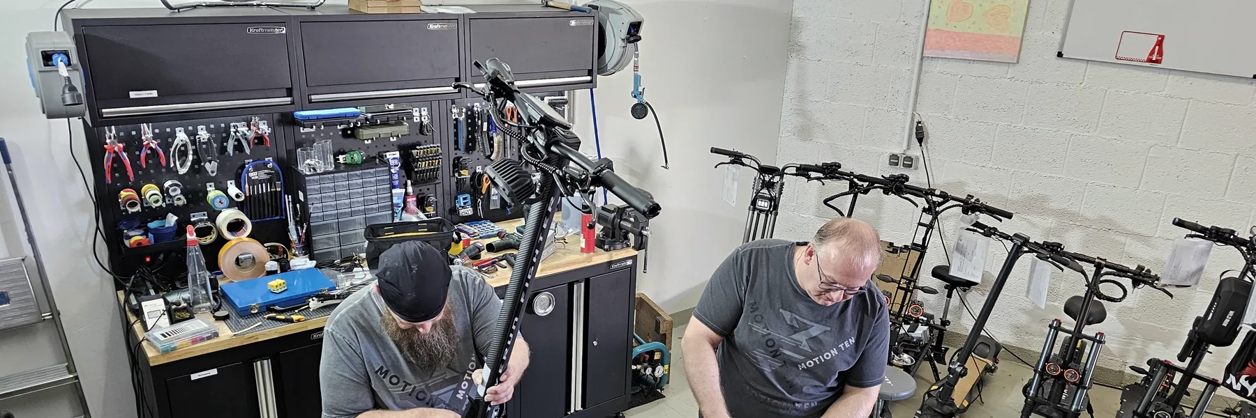 Zwei Techniker arbeiten an Scootern, links die Werkzeugwand, rechts eine Reihe wartender Geräte